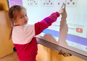 Alicja stoi przed tablicą multimedialną. Dziewczynka wykonała dodawanie i wskazuje kostkę domino z wynikiem.