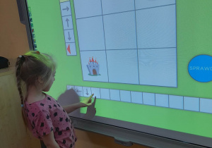 Natalia wyznacza drogę rycerza do zamku na planszy przedstawionej na tablicy multimedialnej. Dziewczynka zapisuje kod za pomocą strzałek.