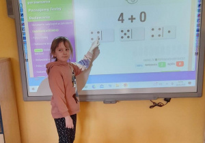 Oliwia podczas gry "Dodawanie w domino" stoi przed tablicą multimedialną. Dziewczynka wykonała dodawanie i wskazuje kostkę domino z wynikiem. Przed tablicą siedzi dwóch chłopców.