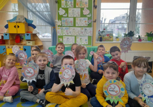 Dzieci siedzą na dywanie i prezentują wykonane przez siebie znaki "Nie pal przy mnie". W tle tablica z pokolorowanymi obrazkami przedstawiającymi postać maskotki programu - Dinka oraz kwiatki, kącik kuchenny, okno.