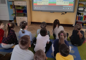 Dzieci siedzą na dywanie przed tablicą multimedialną i oglądają film edukacyjny.