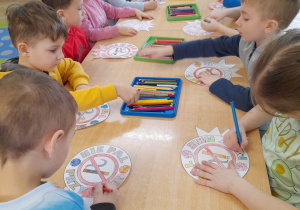 Dzieci siedzą przy złączonych stołach i kolorują znaki "Nie pal przy mnie". Na środku stołów leżą cztery tacki z kredkami.