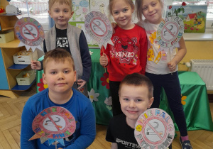 Pięcioro dzieci prezentuje wykonane przez siebie znaki "Nie pal przy mnie". W tle tablica, na której przypięte są pokolorowane przez dzieci obrazki.