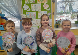 Gabrysia, Alicja, Oliwia i Oskar stoją przed tablicą, na której przypięte są pokolorowane obrazki z maskotką programu - Dinkiem. Przedszkolaki prezentują znaki "Nie pal przy mnie".