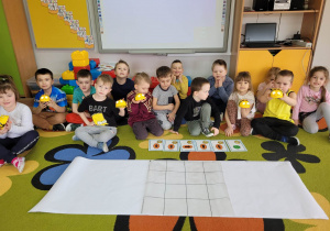 Dzieci z grupy Biedronek siedzą na dywanie z pszczółkami Bee bot w dłoniach. Przed nimi leży plansza 4xc4 oraz zapis kodu na kartach