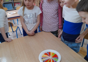 Grupka dzieci stoi przy stole i obserwuje z ciekawością kolorową, cukierkową tęczę na talerzu.