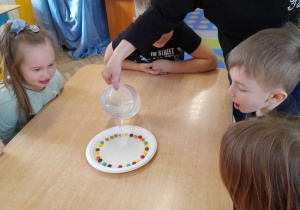 Na środku stołu stoi talerz z kolorowymi cukierkami. Czworo dzieci siedzi przy stole, a Pani Dorotka wlewa wodę na talerz.