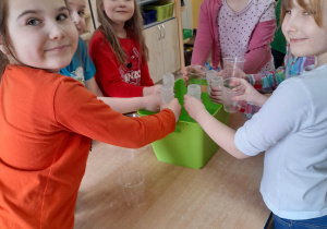 Grupka dzieci stoi przy stole i używając kubków napełnia wodą buteleczki nad pojemnikiem.