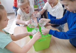 Grupka dzieci nabiera kubkiem wodę z pojemnika i wlewa ją do buteleczek.