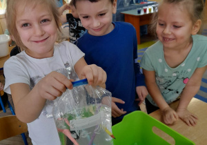 Natalia prezentuje eksperyment "Sprytne ołówki". Dziewczynka trzyma woreczek strunowy z wodą i wbitymi w niego ołówkami. Obok stoi Igor i Gabrysia.