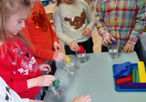 Pięć dziewczynek stoi obok stolika i wykonuje barwniki do eksperymentów. Dziewczynki wkładają kolorową bibułę do kubeczków z wodą.