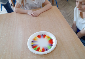 Hubert i Natalia siedzą przy stole podczas zabawy "Cukierkowa tęcza". Na środku stołu stoi talerz z cukierkami i wodą. Dzieci obserwują jak na dnie zaczyna pojawiać się kolorowa, cukierkowa tęcza.