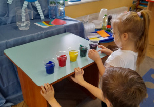 Oskar i Natalia przykucnęli obok stolika, na którym stoją kubeczki z kolorową wodą. Dzieci prowadzą obserwację.