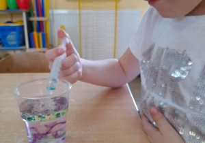 Natalia siedzi przy stole. Dziewczynka podczas zabawy "Perełki w oleju" dodaje strzykawką kolorowe barwniki do kubeczka z wodą i olejem.