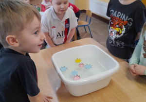 Grupka dzieci stoi przy stole, na którym znajduje się pojemnik z wodą i pływającymi gwiazdkami w kolorze żółtym, różowym i niebieskim. Dzieci obserwują jak gwiazdki rozkładają się na wodzie.