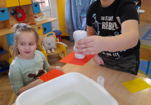 Hubert prezentuje wykonany eksperyment pt. "Czarodziejskie karteczki". Chłopiec trzyma nad miską buteleczkę z wodą odwróconą do góry dnem, pod która znajduje się kartonik w kolorze pomarańczowym. Obok siedzi Alicja.