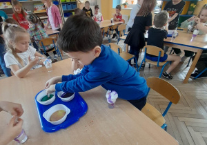 Dzieci siedzą przy stołach i wykonują eksperyment "Kolorowa piana". Przedszkolaki dodają przy użyciu strzykawek kolorowe barwniki na pianę. Przy pierwszym stoliku Igor nabiera zielony barwnik do strzykawki.