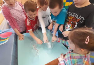 Grupka dzieci stoi przy stole podczas zabawy "Woda wędrowniczka" i sprawdza czy woda przemieszcza się w różnych materiałach.