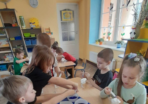 Dzieci siedzą przy dwóch stołach, a przed nimi stoją kubki z wodą i pianką do golenia. Na środku stołu stoją na tacy kolorowe barwniki. Dzieci strzykawkami dodają barwniki na piankę.
