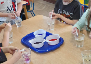 Na stole stoi tacka z barwnikami, a przed dziećmi znajdują się kubki z wodą i olejem. Grupka dzieci podczas zabawy "Perełki w oleju" dodaje strzykawką kolorowe barwniki do kubka i obserwuje jak powstają kolorowe perełki i jak się przemieszczają.
