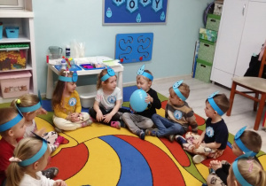Dzieci siedzą na dywanie w kole i podają sobie niebieski balon wypełniony wodą. Przedszkolaki mają założone na głowach niebieskie opaski z kropelkami. W tle dekoracja z okazji Dnia Wody.