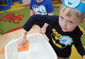 Hubert podczas zabawy badawczej "Czarodziejskie kartoniki". Chłopiec nad miską trzyma do góry dnem białą buteleczkę wypełnioną wodą i zakrytą pomarańczową karteczką.