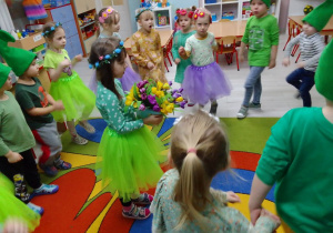Dziewczynki w kolorowych tiulowych spódniczkach z kwiatkami na głowie, chłopcy w zielonych czapkach na głowach poruszają się w kole do muzyki, w środku Natalia z bukietem krokusów.