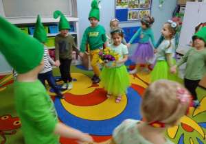 Dzieci w wiosennych przebraniach poruszają się w kole trzymając się za ręce. W środku koła Matylda, która trzyma bukiet z kolorowymi krokusami.