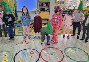 Dzieci z grupy "Biedronek" wykonują wiosenne zadanie matematyczne. Na podłodze obok dzieci widać rozłożone hula hop, kolorowe kwiatki i kartoniki z cyferkami.