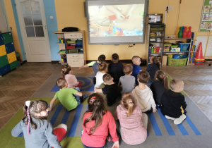 "Słoneczka" siedzą na dywanie przed tablicą multimedialną i oglądają film przedstawiający wykonanie Marzanny.
