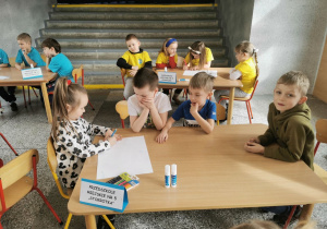 Ala siedzi przy stoliku i rysuje na kartce zgodnie z instrukcją pani prowadzącej konkurs. Chłopcy podpowiadają dając wskazówki co i gdzie ma rysować Ala.