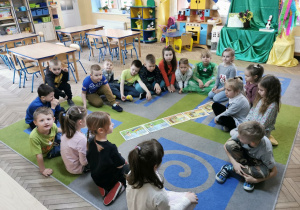 "Słoneczka" siedzą na dywanie w kole i słuchają opowiadania czytanego przez nauczyciela. Na środku widać kolorowe ilustracje