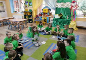 Dzieci z grupy "Słoneczka" siedzą w kole na dywanie. Na środku siedzi Vanessa, która trzyma kartę, na której wskazuje klamerką cyfrę odpowiadającą ilości bocianów.