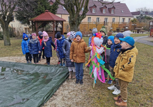 Dzieci z grupy "Słoneczka" stoją przed piaskownicą wypowiadając słowa rymowanki. Krzysio i Oliwier trzymają Marzannę.