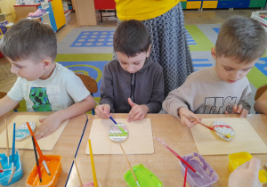 Karol, Igor i Oskar siedzą przy stole i malują kolorowym lukrem pisankowe ciasteczka.