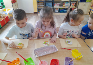Mieszko, Alicja, Alicja i Bruno siedzą obok siebie przy stole i malują kolorowym lukrem pisankowe ciasteczka.