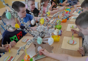 Dzieci siedzą przy złączonych stołach i ozdabiają pisankowe ciasteczka. Na środku stołów stoją w pojemnikach kolorowe lukry, wazon z baziami i pisankami oraz drewniany zając.