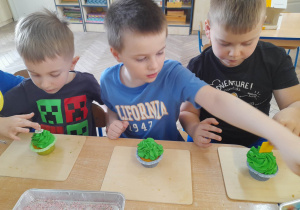Oliwier, Krzyś i Hubert siedzą przy stole, a przed nimi na tackach leżą muffinki z zielonym kremem.