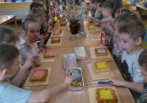 Dzieci siedzą przy złączonych stołach, a przed nimi na deseczkach leżą mazurki. Na środku stołów znajdują się w pojemnikach cukiereczki, posypki, lukier do dekoracji mazurków.