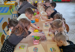 Dzieci siedzą przy złączonych stołach i ozdabiają pisankowe ciasteczka lukrem, posypkami.