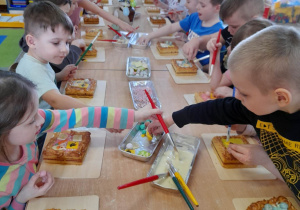 Dzieci siedzą przy złączonych stołach, a przed nimi leżą na deseczkach mazurki. Przedszkolaki kończą dekorowanie ciasta.