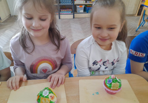 Alicja i Alicja siedzą obok siebie przy stole i z radością prezentują udekorowane przez siebie muffinki.