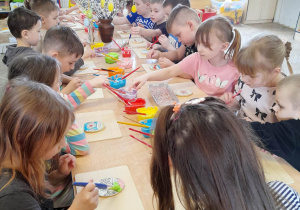 Dzieci siedzą przy złączonych stołach i malują kolorowym lukrem przy użyciu pędzli pisankowe ciasteczka. W tle tablica z Kodeksem Przedszkolaka, kącik sportowy, muzyczny, okno.