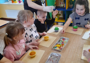 Czworo dzieci siedzi przy stole, a obok stoi Pani, która pomaga dzieciom wycisnąć zielony krem na muffinkę. Wiktoria rozpoczęła dekorowanie swojej muffinki.