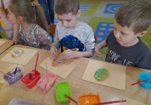 Vanessa, Hubert i Tomek siedzą przy stole, a przed nimi leżą deseczki z ciasteczkami i lukry w pojemnikach. Dzieci dekorują pisankowe ciastko kolorowymi posypkami.