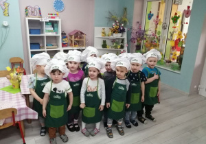Zdjęcie grupowe dzieci. Przedszkolaki ubrane są w zielone fartuszki i białe czapki kucharskie. W tle szafki z układankami i kącik przyrodniczy.