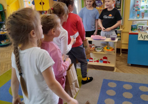 Dzieci bawią się w kąciku tematycznym "Sklep". Bruno i Alicja stoją za ladą, a cztery dziewczynki z torbami w ręku czekają w kolejce.