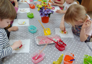 Dzieci z grupy "Motylków" przy stolikach ozdabiają pędzelkami i kolorowym lukrem ciastka w kształcie jajka. Na stoliku są pojemniki z kolorowym lukrem, pędzle, kolorowe posypki i wiosenny stroik.