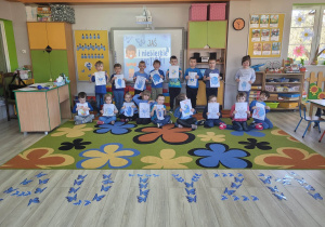 Dzieci z grupy Biedronek stoją na dywanie przed tablicą multimedialną. Każde dziecko trzyma w dłoniach wykonaną przez siebie pracę plastyczną zatytułowaną „Dziś bądź niebieski”. Przed dziećmi na podłodze widać ułożony z niebieskim motylków napis „Autyzm”.