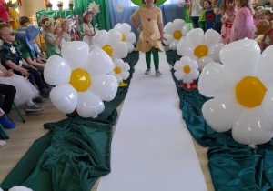 Ala z grupy "Biedronki" idzie po białym dywanie między balonami w kształcie stokrotek. Po bokach siedzą dzieci, a w tyle znajduje się dekoracja z okazji Dnia Ziemi.